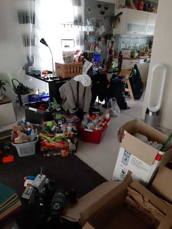 Unaufgeräumtes Wohnzimmer mit Kartons, Spielsachen, Haushaltswaren und Unordnung.