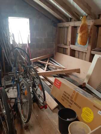 Dachboden mit alten Fahrrädern, Holzlatten, Kartons und hängendem Schinken bei Tageslicht.